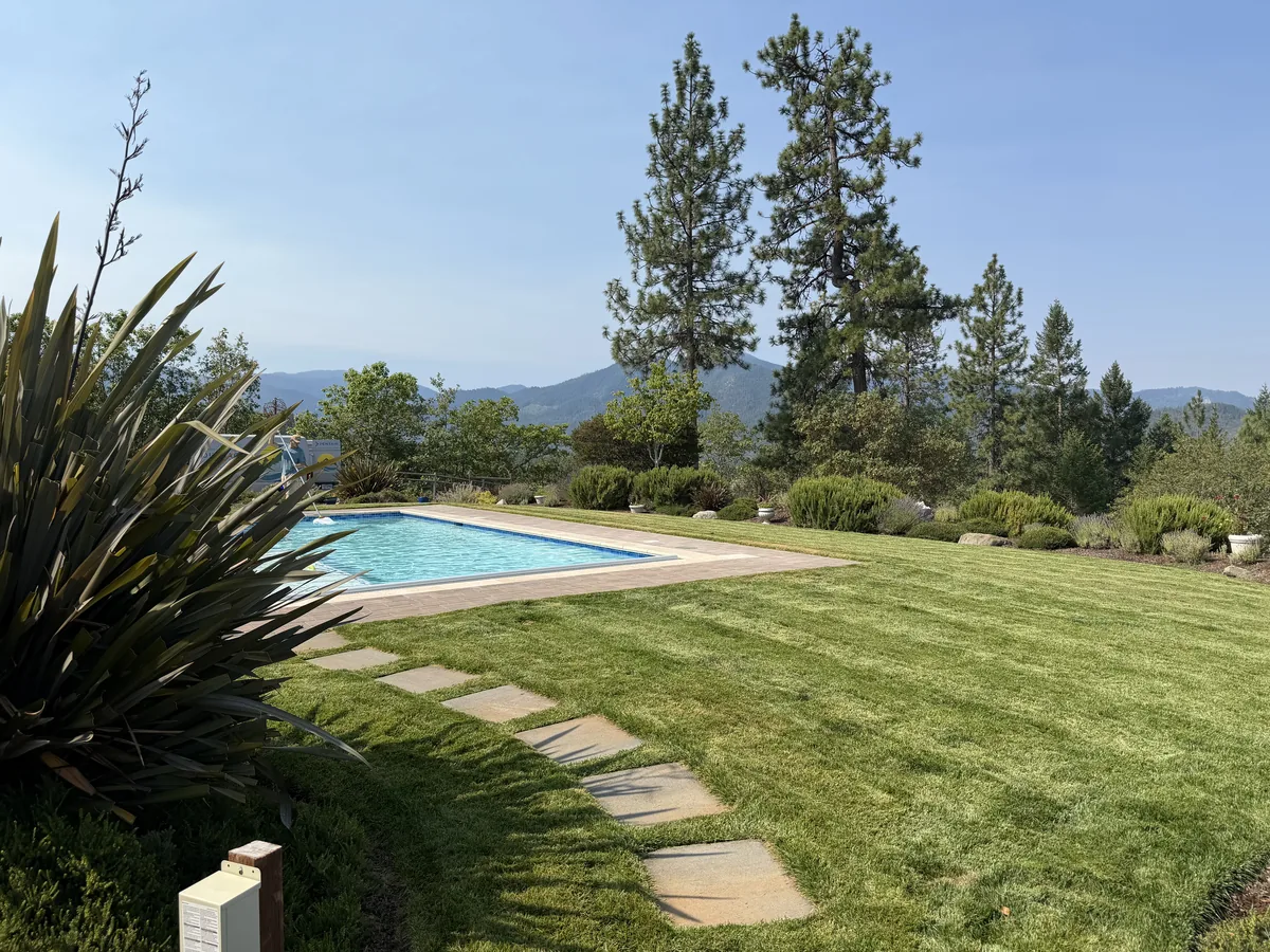 Manicured lawn, flagstone stepping stone path and trimmed shrub beds around a backyard pool with Southern Oregon hills behind